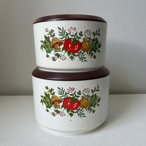 May include: Two stacked cylindrical white storage containers with dark brown lids. The exterior features a colorful botanical print of red tomatoes, yellow onions, green peppers, mushrooms, and small red berries surrounded by green leaves and stems.