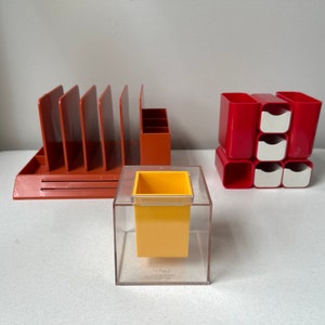 May include: A collection of retro office storage solutions in orange, red, and yellow. The image shows a file organiser, a pen holder, a modular storage unit, and a small yellow container in a clear acrylic cube. Perfect for desk organisation.