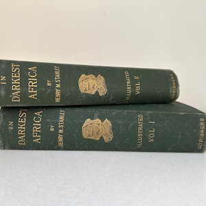 May include: Two vintage books titled "In Darkest Africa" by Henry M. Stanley. The books are dark green with gold lettering and illustrations. Volume I and Volume II are stacked on top of each other.