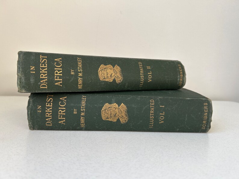 May include: Two vintage books titled "In Darkest Africa" by Henry M. Stanley. The books are dark green with gold lettering and illustrations. Volume I and Volume II are stacked on top of each other.