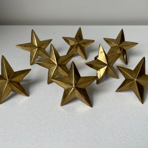 May include: A collection of eight brass star-shaped decorative objects. Each star has five points and a slightly aged, golden finish. The stars are arranged on a white surface, showcasing their dimensional design.