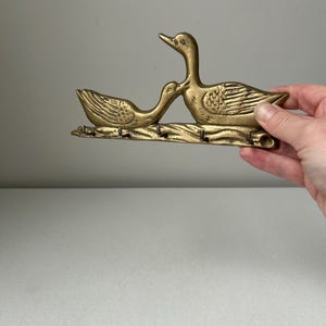 May include: A brass wall-mounted key holder featuring two ducks facing each other. The ducks are positioned above a horizontal bar with five small metal hooks extending downward for hanging items. The metal has a polished, golden-toned finish.