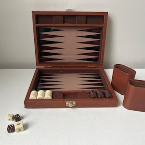 May include: A brown backgammon set featuring a folding case with a dark brown and tan triangular point board, two brown leather dice cups, white and dark brown checkers, and four dice, including one with the number two clearly visible.