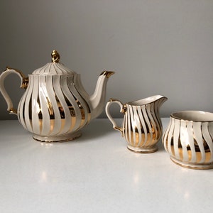 May include: A tea set consisting of a teapot, a creamer, and a sugar bowl. Each piece is made of off-white ceramic featuring vertical, swirling gold metallic stripes. The teapot has a lid with a gold finial, a curved handle, and a spout.