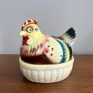 May include: A ceramic hen-shaped container with a yellow, brown, and green color scheme. The hen sits atop a white basket-like base. The hen has a red comb and beak, and a blue eye. A decorative kitchen item.