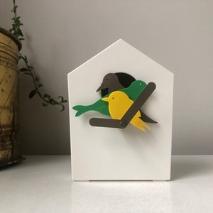 May include: A white, birdhouse-shaped clock with a brown, green, and yellow bird design. The clock features a dark brown branch and a black clock face. The birds are perched on a branch, adding a whimsical touch to the design.