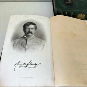 May include: Open book with a black and white portrait of a man with a large mustache and signature. The book is open to a page with the portrait and text. A green book with gold lettering is visible in the background.
