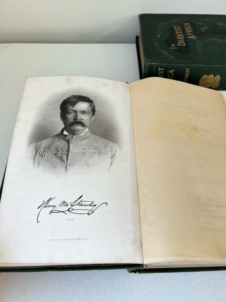 May include: Open book with a black and white portrait of a man with a large mustache and signature. The book is open to a page with the portrait and text. A green book with gold lettering is visible in the background.