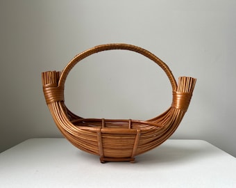 Könnte beinhalten: Ein handgefertigter, oval geformter Korb aus geflochtenem Rattan. Der Korb hat einen großen, bogenförmigen Griff und einen stabilen Boden. Die natürliche braune Farbe des Rattans verleiht ihm ein rustikales und organisches Aussehen. Dieser dekorative Korb ist für verschiedene Zwecke geeignet.