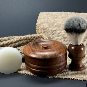 May include: A wooden shaving set with a bowl, soap, and brush. The bowl and brush are made of dark wood, and the soap is white. The set is on a burlap background.