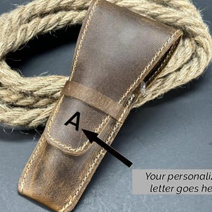 May include: A brown leather case with a stitched edge and a letter "A" embossed on the front. The case is designed to hold a straight razor.