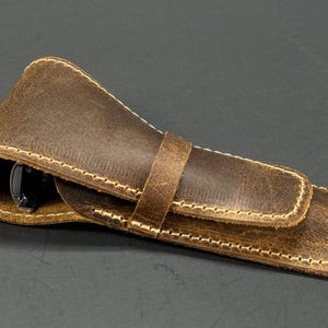 May include: A brown leather pen case with a flap closure and a stitched design. The case is made of distressed leather and has a light brown stitching.