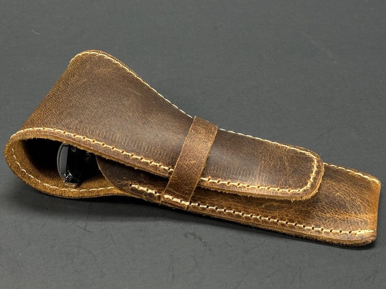 May include: A brown leather pen case with a flap closure and a stitched design. The case is made of distressed leather and has a light brown stitching.