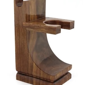 Puede incluir: Un soporte de madera marrón oscuro para brocha de afeitar y maquinilla. El soporte tiene un diseño curvo y presenta un soporte circular para una brocha de afeitar y una ranura para una maquinilla. La base es rectangular.