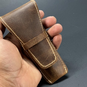 May include: A brown leather case with a stitched design and a flap closure. The case is held in a hand.