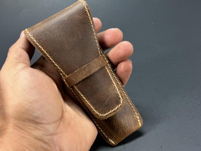 May include: A brown leather case with a stitched design and a flap closure. The case is held in a hand.