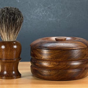 May include: A wooden shaving brush with a dark brown handle and a brush head with light and dark bristles. Next to it is a matching wooden shaving soap bowl with a lid. The items are made of dark wood.