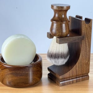 Puede incluir: Un soporte de madera para brocha de afeitar con una brocha de afeitar blanca, un jabonera de madera con una pastilla de jabón blanca y una base de madera.