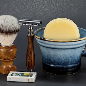 Puede incluir: Un juego de afeitado con una brocha de afeitar con mango de madera, una maquinilla de afeitar de seguridad con mango de madera, un cuenco de afeitar de cerámica azul con jabón y una pequeña caja de cuchillas. Sobre una superficie gris oscuro.