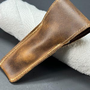 May include: A brown leather case with a stitched edge. The case is lying on a white towel.