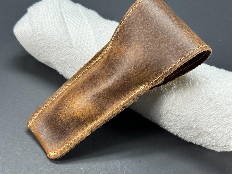May include: A brown leather case with a stitched edge. The case is lying on a white towel.