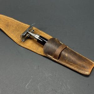 May include: A brown leather case with a stitched edge holds a silver and black safety razor.