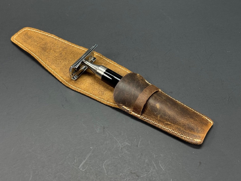 May include: A brown leather case with a stitched edge holds a silver and black safety razor.