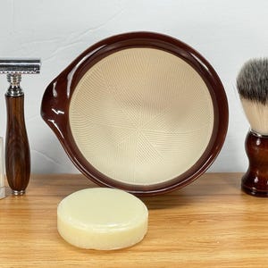 Puede incluir: Un juego de afeitado con una maquinilla de afeitar de seguridad con mango marrón, una brocha de afeitar, un cuenco de afeitar y una pastilla de jabón. El cuenco es marrón con el interior crema. El cepillo tiene un mango de madera oscura y una cabeza de cerdas grises y blancas.