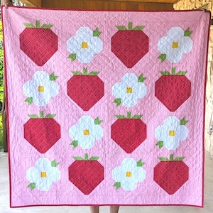 May include: A square quilt featuring a repeating pattern of red strawberries and white flowers with yellow centers on a light pink background. The quilt has a red and white polka dot border and is stitched with a diamond-shaped quilting pattern.