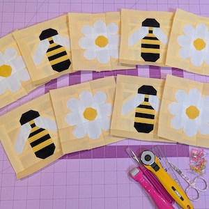 Op de afbeelding: Een verzameling gele stoffen vierkanten met applicaties van bijen en madeliefjes. De bijen zijn zwart en geel, en de madeliefjes zijn wit met gele centra. Naaigerei, waaronder een rolmes en een schaar, zijn ook zichtbaar.