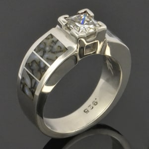 Op de afbeelding: Een zilveren ring met een vierkante, heldere edelsteen in een kraalbezetting. De band is versierd met een patroon van gefossiliseerd dinosaurusbot.