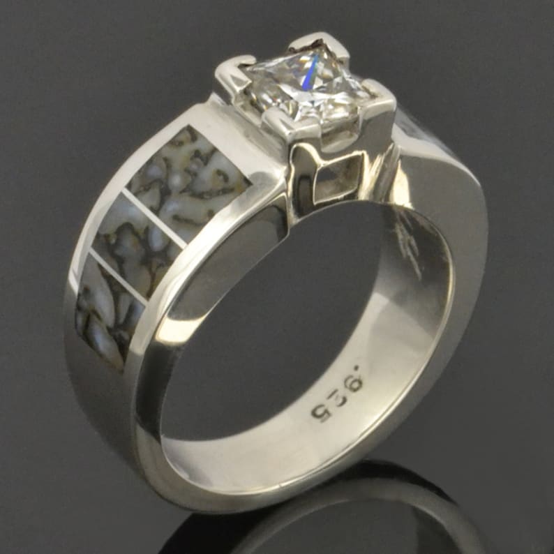 Op de afbeelding: Een zilveren ring met een vierkante, heldere edelsteen in een kraalbezetting. De band is versierd met een patroon van gefossiliseerd dinosaurusbot.