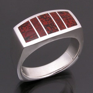 May include: A silver ring with a rectangular band featuring five inlaid red and black fossilized dinosaur bone pieces.