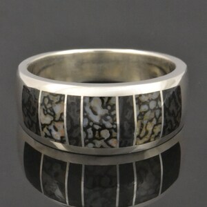 May include: A silver band ring with a black and white fossilized dinosaur bone inlay. The ring has a polished finish and is set with a row of black and white fossilized dinosaur bone inlay.