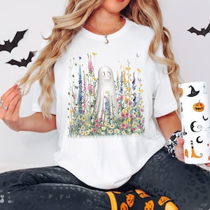 Puede incluir: Camiseta blanca con un diseño de fantasma sonriente en un campo de flores silvestres coloridas. El diseño tiene un tema caprichoso de Halloween. Un vaso blanco con ilustraciones de Halloween se sostiene en la mano.