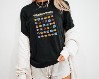 Op de afbeelding: Zwart T-shirt met een afbeelding van kleurrijke bloemen en de tekst "Grow Positive Thoughts, Grow Beautiful Things".