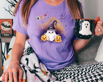 Può includere: Una maglietta lilla con un disegno di fantasma di Halloween, una tazza in bianco e nero con un fantasma e una coperta a tema Halloween. La maglietta presenta un fantasma con un cappello da strega, zucche e la parola "BOO".