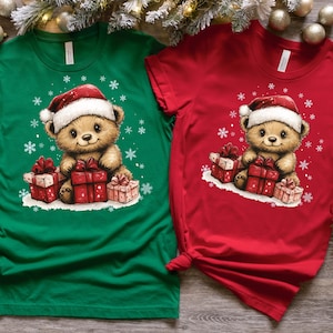 May include: Two Christmas themed t-shirts featuring a cartoon teddy bear wearing a Santa hat and holding presents. One shirt is green and the other is red.