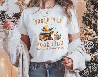 Könnte beinhalten: Weißes T-Shirt mit einem Grafikdesign aus einem Stapel Bücher, einer Tasse heißer Schokolade und einer brennenden Kerze. Der Text "North Pole Book Club Hot Cocoa Served Daily" ist auf das Shirt gedruckt.