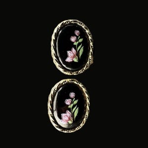 May include: A pair of oval-shaped earrings. Each earring features a black background with a painted pink flower and green leaves. The earrings are framed by a gold-colored, rope-like border. The earrings are displayed against a black background.