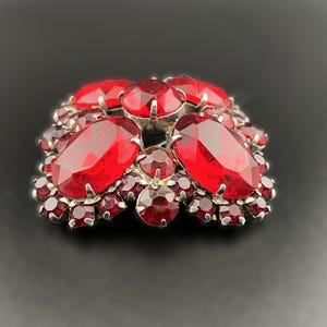 May include: A vintage brooch featuring a cluster of faceted, deep red gemstones. The brooch has a silver-toned metal setting and is composed of various sizes of stones, creating a sparkling, eye-catching design. The brooch is a classic piece of jewelry.