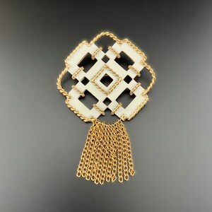 May include: A vintage brooch with a geometric design. The brooch features a cream-colored, square-shaped center with a gold-colored, braided border. A gold-colored chain tassel hangs from the bottom. The brooch is set against a dark background.