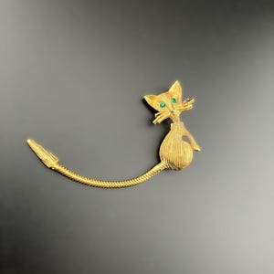 May include: A gold-toned cat brooch with green gemstone eyes. The cat has a long, curved tail and a textured body. The brooch is set against a dark background.
