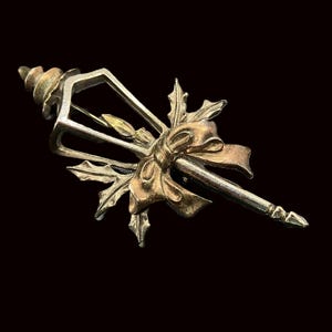 May include: A vintage brooch with a gold and silver design. The brooch features a bow, holly leaves, and a decorative element. The pin is set against a black background, highlighting the intricate details and metallic colors.