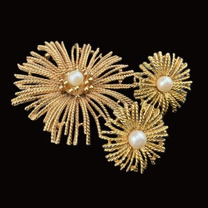 May include: A gold-toned floral brooch and matching earrings set against a black background. The brooch features three stylized flower designs with textured, radiating petals and a central pearl. The earrings mirror the smaller flower design.