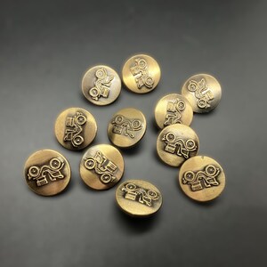 May include: A collection of ten gold-toned metal buttons with a raised design of a vintage car. The buttons are scattered on a gray surface.
