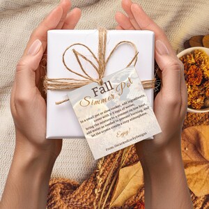 Może przedstawiać: Białe pudełko prezentowe związane sznurkiem, z etykietą z napisem "Fall Simmer Pot". Etykieta zawiera instrukcje użytkowania. W tle kubek wypełniony suszonymi kwiatami i jesiennymi liśćmi, tworzący przytulny jesienny motyw.