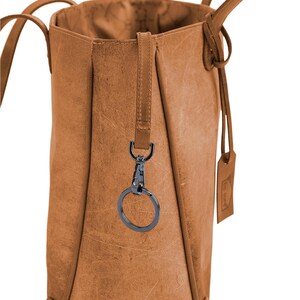 K&ouml;nnte beinhalten: Eine braune Ledertasche mit einem schwarzen Schl&uuml;sselring aus Metall, der an der Seite befestigt ist. Die Tasche hat einen langen Riemen und ein kleines Etikett mit dem Wort "Leder" darauf.