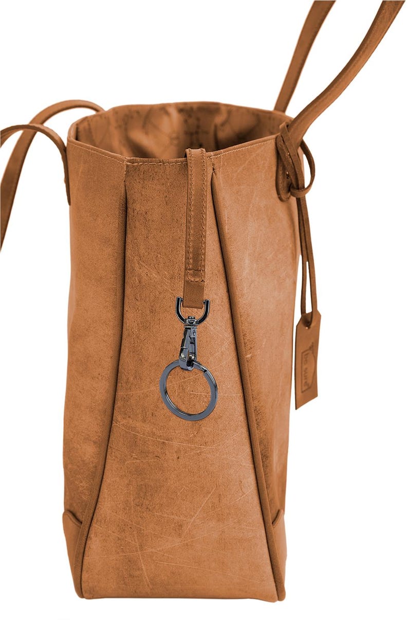 K&ouml;nnte beinhalten: Eine braune Ledertasche mit einem schwarzen Schl&uuml;sselring aus Metall, der an der Seite befestigt ist. Die Tasche hat einen langen Riemen und ein kleines Etikett mit dem Wort "Leder" darauf.