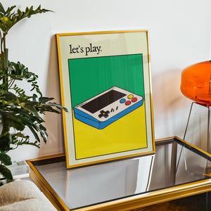 May include: A framed poster with a yellow, green, and blue color scheme. The poster features a cartoon illustration of a handheld video game console with the text "Let's play."